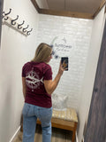 Person wearing a maroon shirt with an image, standing in a room with a wooden floor and white walls.